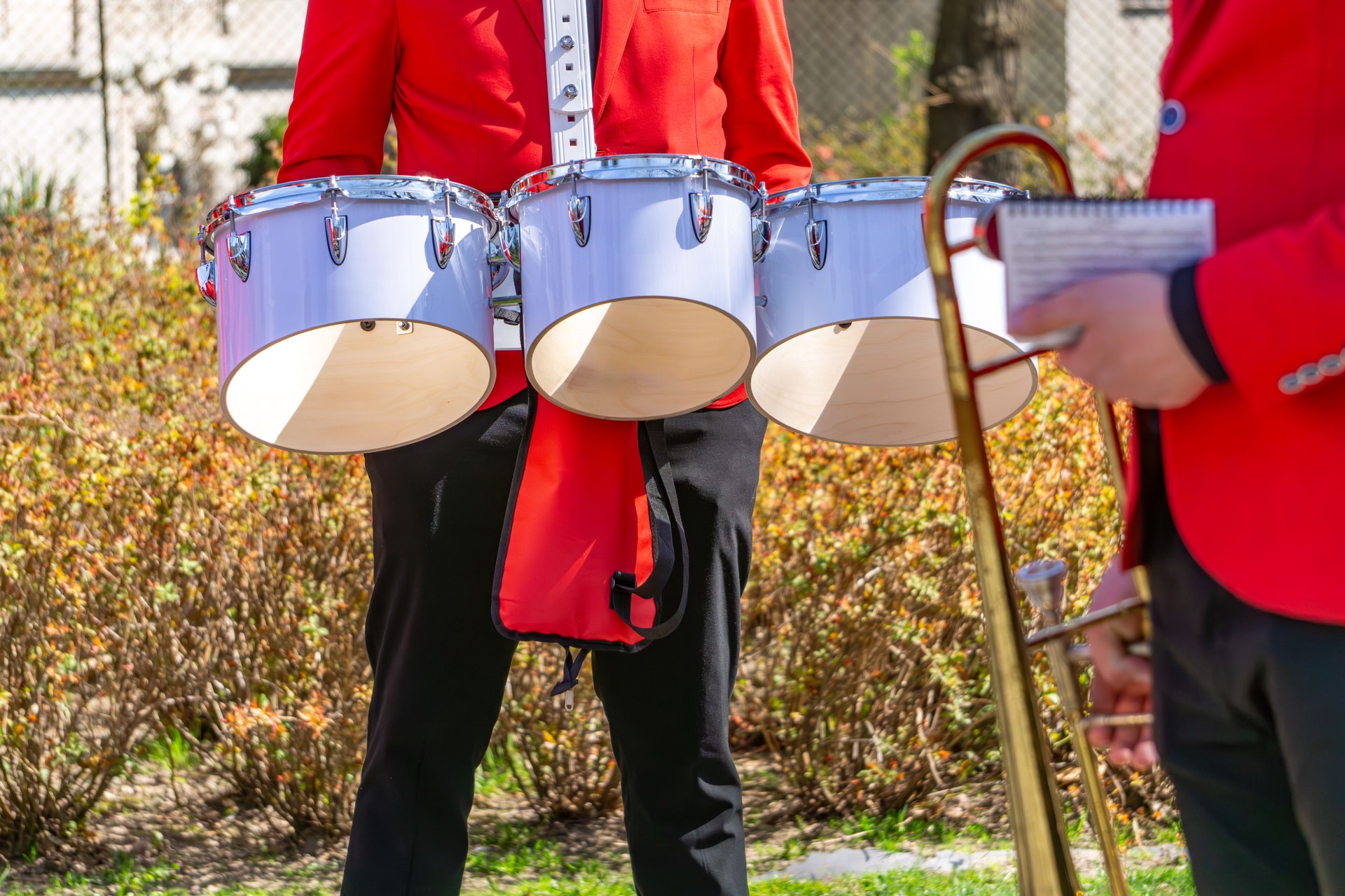 Musicians in red uniforms playing wind and percussion instruments during an outdoor performance. In the foreground are snare drums worn on harnesses.